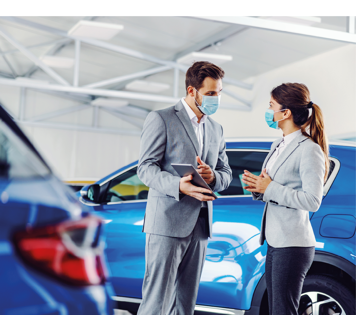 Car seller standing in car salon with a customer and showing on tablet car performances and specifications  They both have face masks on faces because it's corona outbreak 