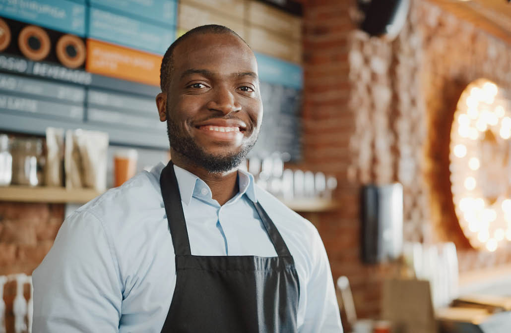 Handsome Black African American Barista with Short Hair and Beard Wearing Apron is Smiling in Coffee Shop Restaurant  Portrait of Happy Employee Behind Cozy Loft-Style Cafe Counter 