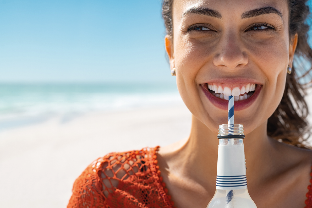 Closeup face of young woman drinking fresh sparkling water from a glass bottle at beach. Portrait of beautiful carefree girl drinking soft drink with straw during summer. Smiling girl staying hydrated