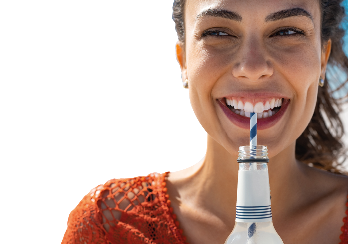 Closeup face of young woman drinking fresh sparkling water from a glass bottle at beach. Portrait of beautiful carefree girl drinking soft drink with straw during summer. Smiling girl staying hydrated