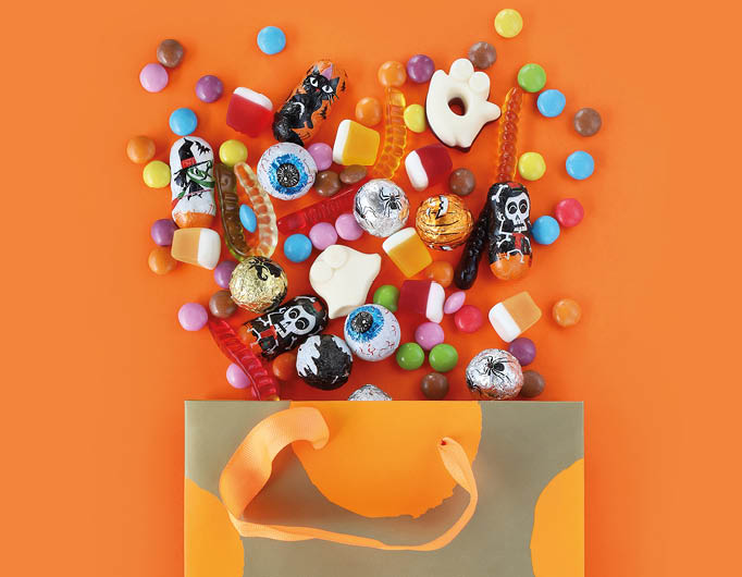 Shopping paper orange polka dot gift bag full of spilled assorted traditional Halloween candies. Orange banner background with copy space. Happy Halloween holiday sale and trick or treat concept.