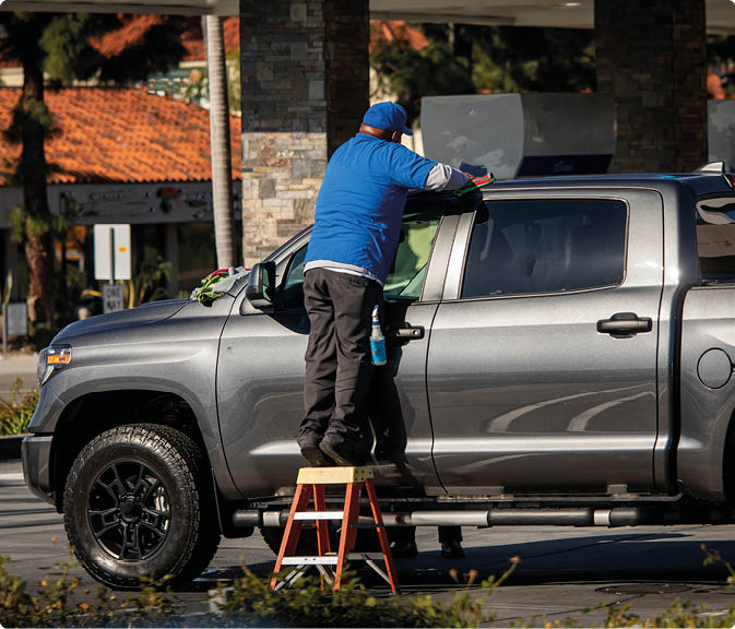 Car wash worker standing on small ladder cleaning a light truck in bright sun. Horizontal image