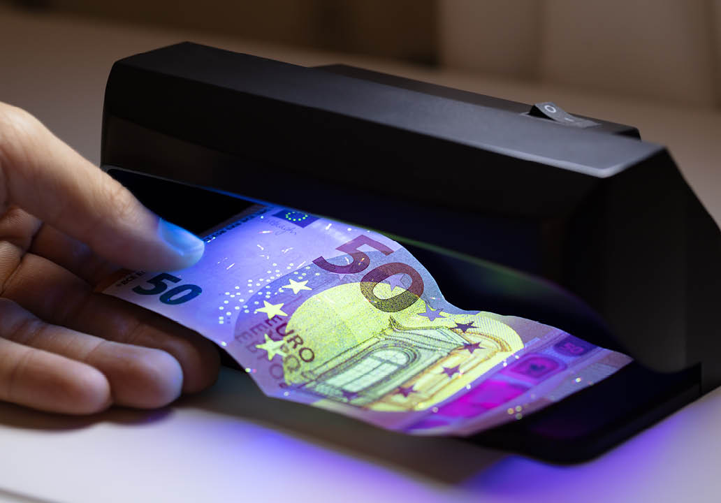 Close-up Of Man Hand Checking Bank Note Through Currency Detector Technology On White Desk