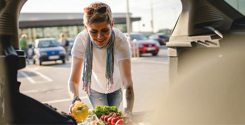 One woman mature caucasian female putting box with vegetables in the back of her car in sunny day on the parking lot in front of supermarket or grocery store in sunny day copy space real people