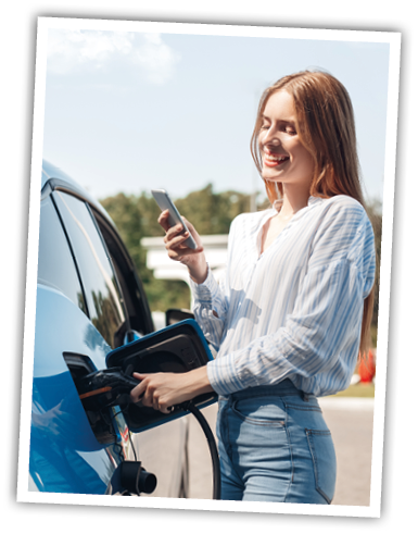 Young beautiful woman traveling by electric car having stop at charging station standing plugging cable browsing internet on smartphone smiling joyful while charing
