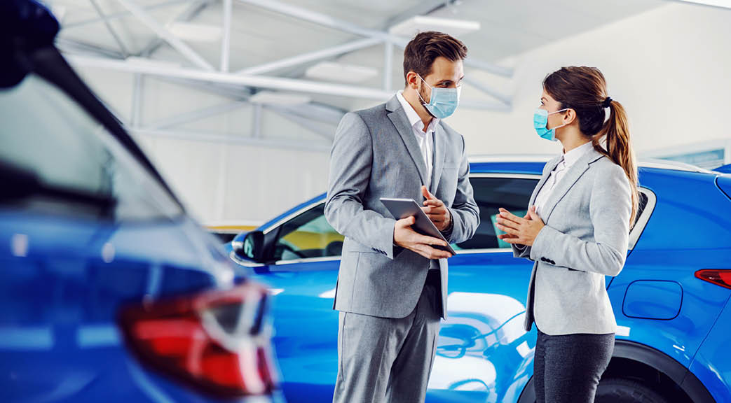 Car seller standing in car salon with a customer and showing on tablet car performances and specifications  They both have face masks on faces because it's corona outbreak 