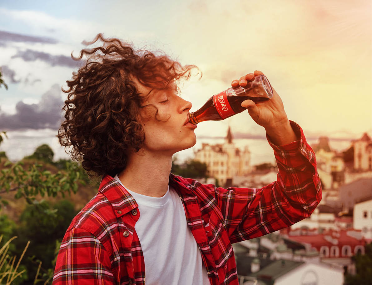 Kyiv, Ukraine, August 1, 2020  Handsome young man with curly hair and in a red plaid shirt holds a glass bottle of Coca-Cola in his hand and drinks from it 