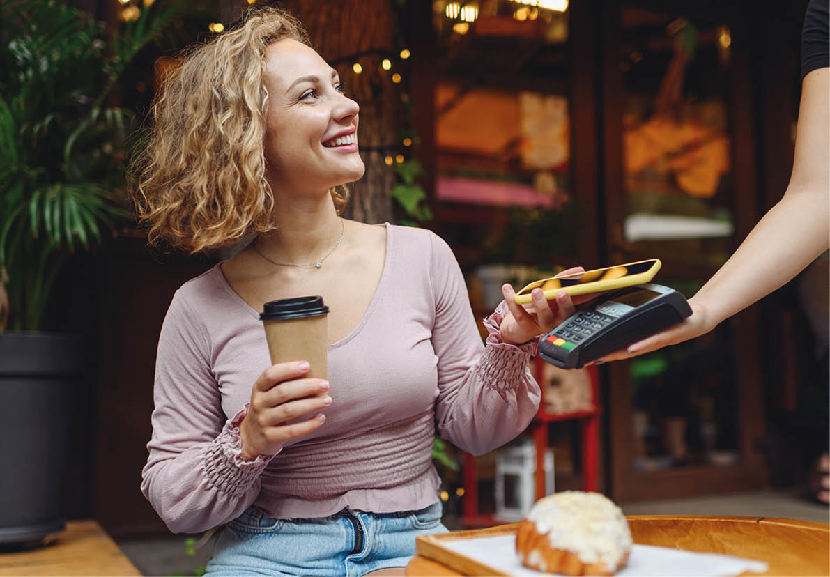 Young smiling woman 20s in casual clothes at cafe buy breakfast sit at table hold wireless bank payment terminal mobile phone to process acquire payments relax in restaurant during free time indoors.