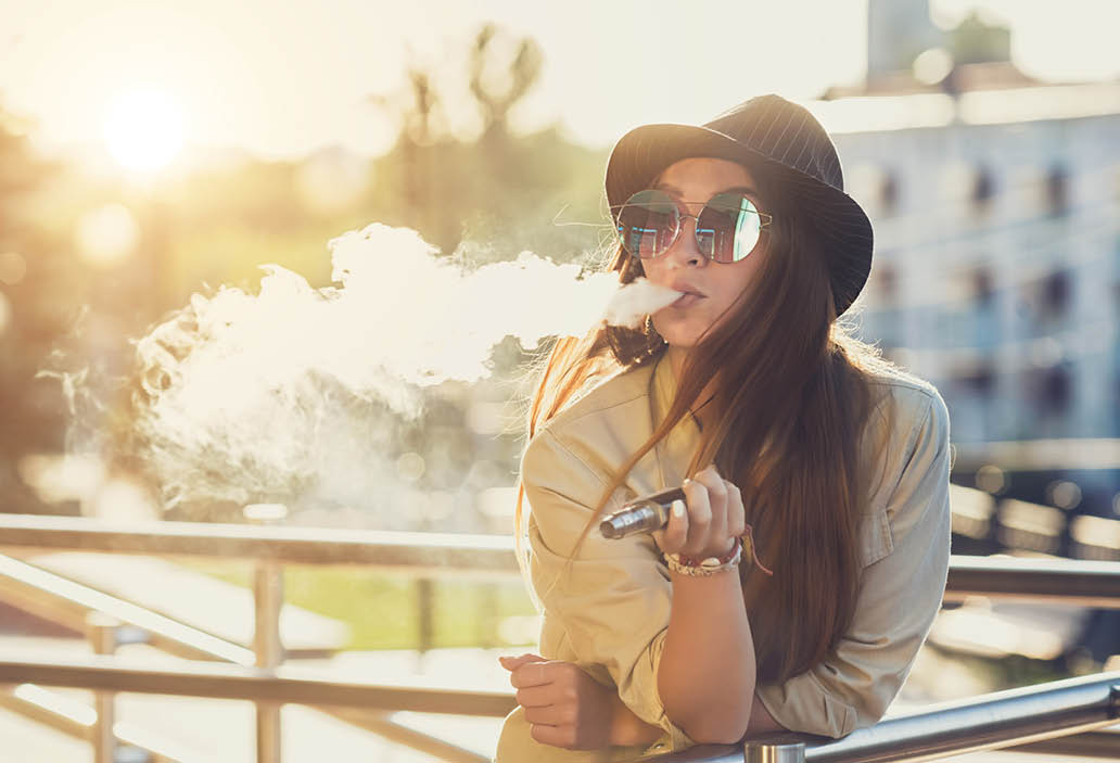 Pretty young hipster woman in black hat vape ecig, vaping device at the sunset. Toned image