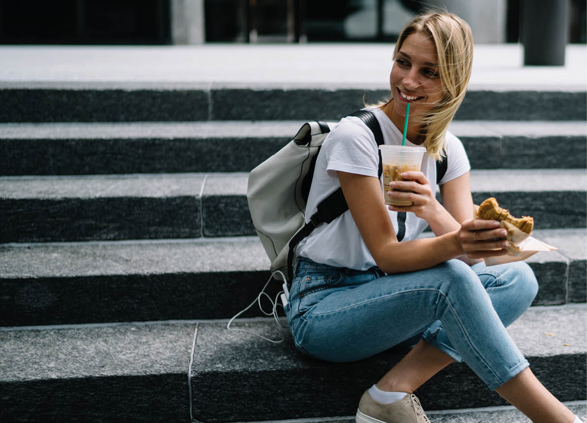 Smiling hipster girl resting on stairs enjoying eating street food on lunch, happy female traveler tasting snack and drinking ice coffee sitting on urban setting having break during walking city trip 