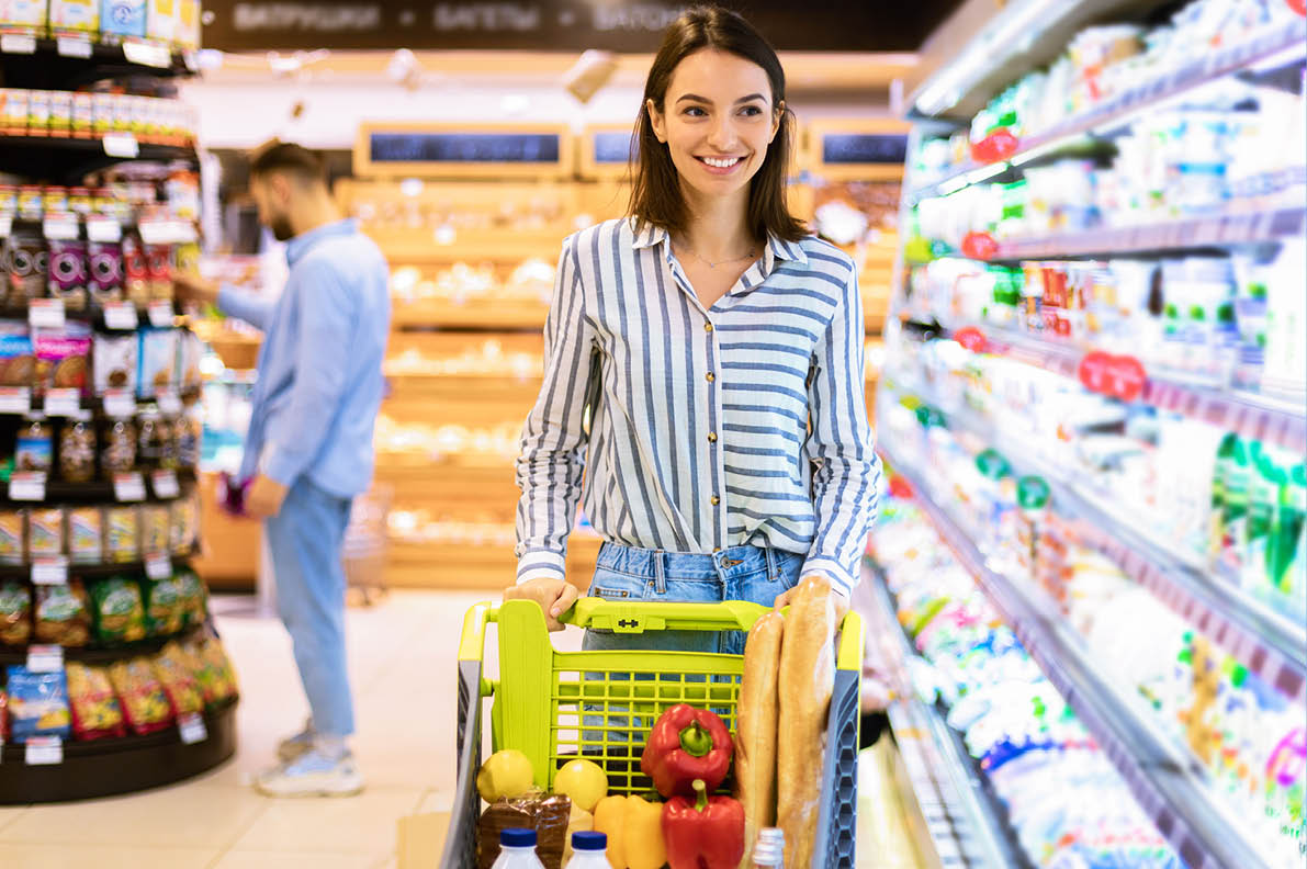 Smiling Cheerful Young Woman Walking With Shopping Trolley Cart Along The Shelves In Grocery Store  Happy Female Customer Buying Groceries In Supermarket, Looking At Fridge With Dairy Products
