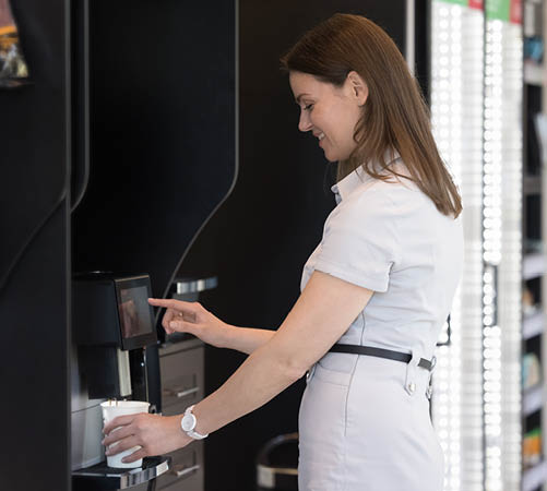Young attractive businesswoman prepare morning hot beverage using professional vending machine, standing alone in modern office workspace during lunch coffee break. Food and drink industry, lifestyle