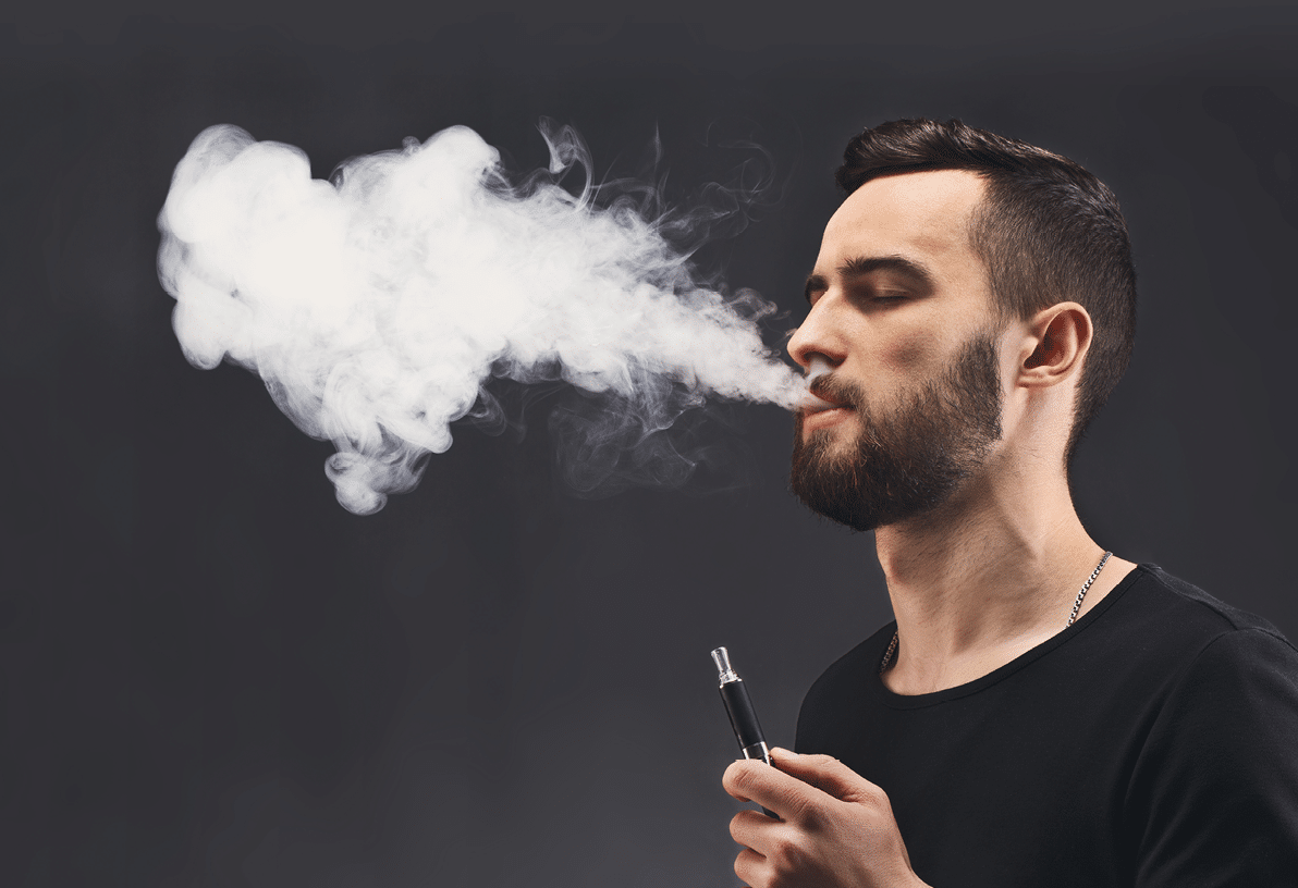 Young man vaping, studio shot. Bearded guy blowing a heart-shape cloud of smoke on black background. Nicotine free smoking and vapor concept, copy space.