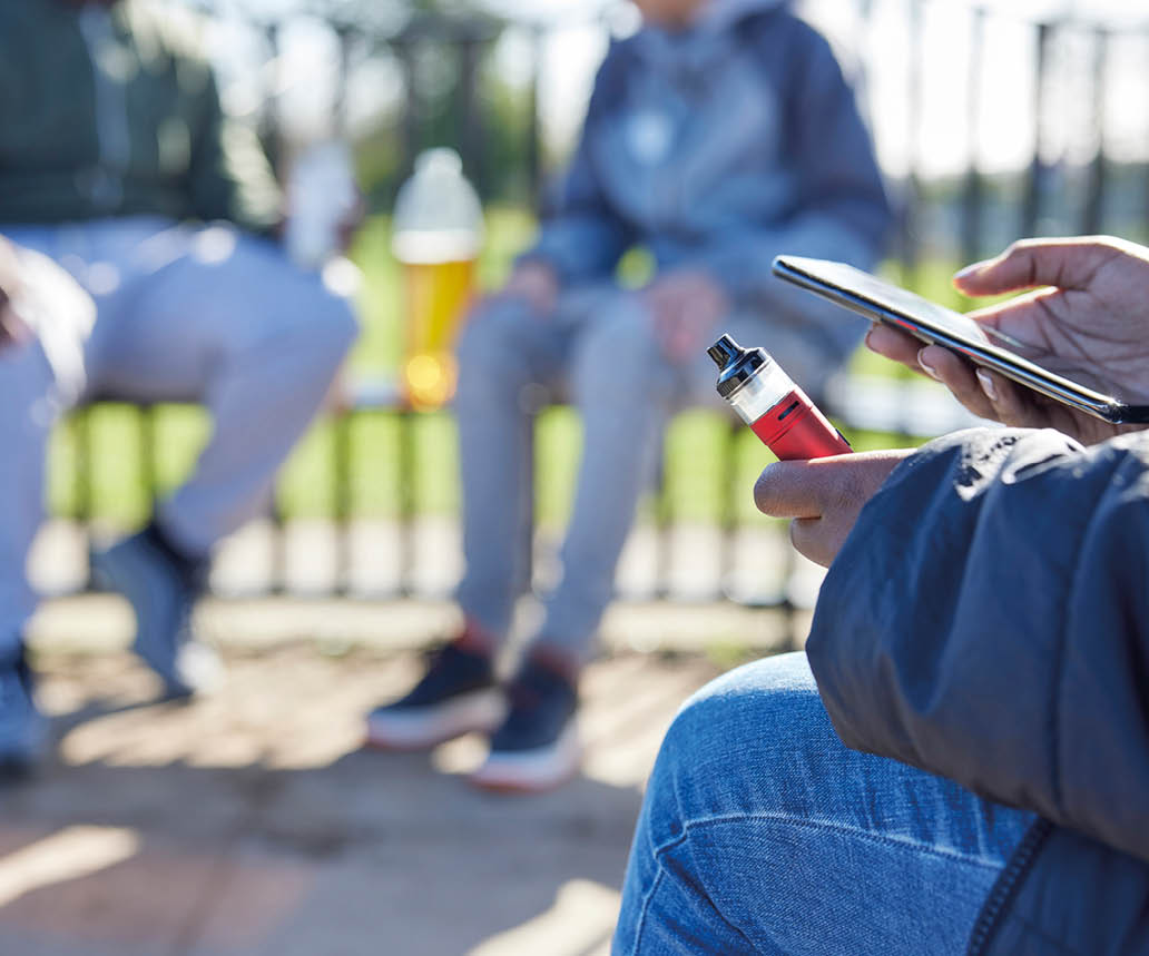 Close Up Of Teenagers With Mobile Phone Vaping and Drinking Alcohol In Park