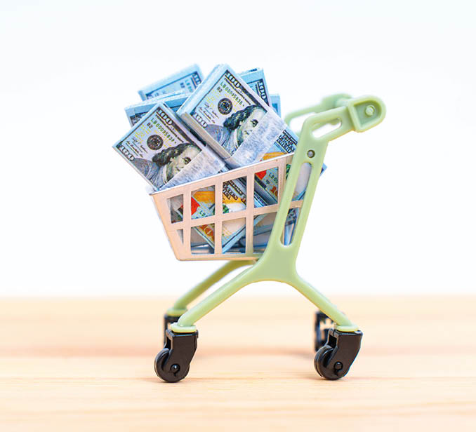 Mini shopping cart overflowing with toy currency on a wooden background, representing concepts of finance, shopping, and consumerism