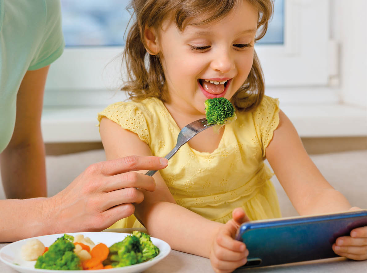 A happy child eats broccoli only with cartoons on his phone. Organic cabbage and food on a plate. The problem of feeding the children preschool age