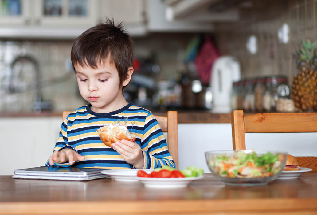 Beautiful little boy, eating sandwich at home, playing on tablet, vegetables on the table