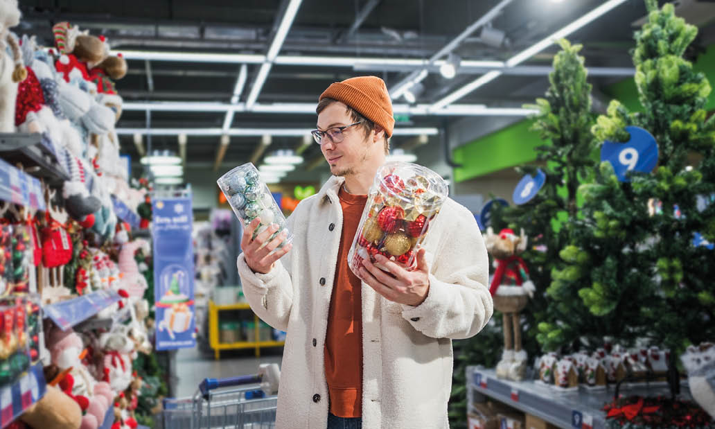 A man in a supermarket chooses Christmas tree balls and decorations. A stylish guy in a white fur coat looks at Christmas decorations on the shelves in the store, festive mood, buying Christmas gifts