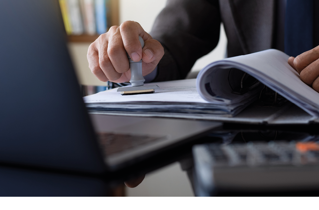 Businessman in suit hand stamping rubber stamp on document in file folder with laptop computer on the desk at office. Authorized allowance permission approval concept.