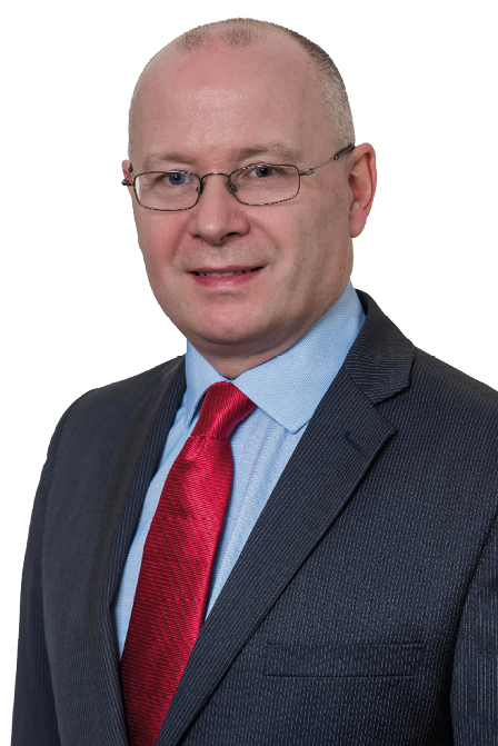 A person wearing glasses, a dark suit, light blue shirt, and red tie is smiling against a white background. Description generated by AI