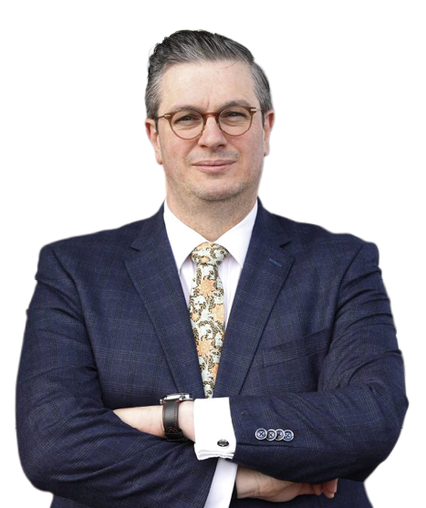 A person with short slicked-back hair and glasses is wearing a dark blue suit with a white shirt and a patterned tie while standing with arms crossed against a white background. Description generated by AI