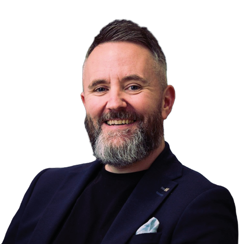 A person with short hair and a beard is smiling and wearing a dark blazer over a black shirt with a light-colored pocket square. Description generated by AI