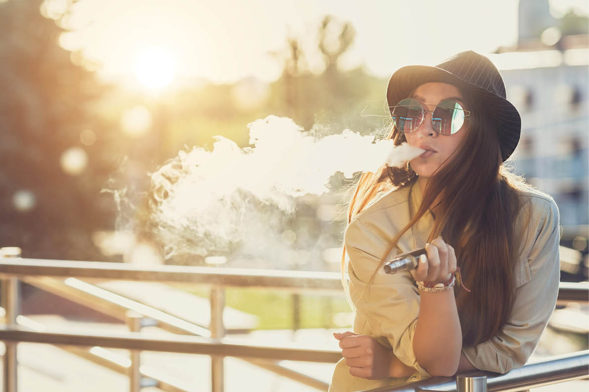 A person wearing sunglasses and a hat is leaning on a railing outdoors while exhaling vapor from an electronic cigarette during sunset. Description generated by AI
