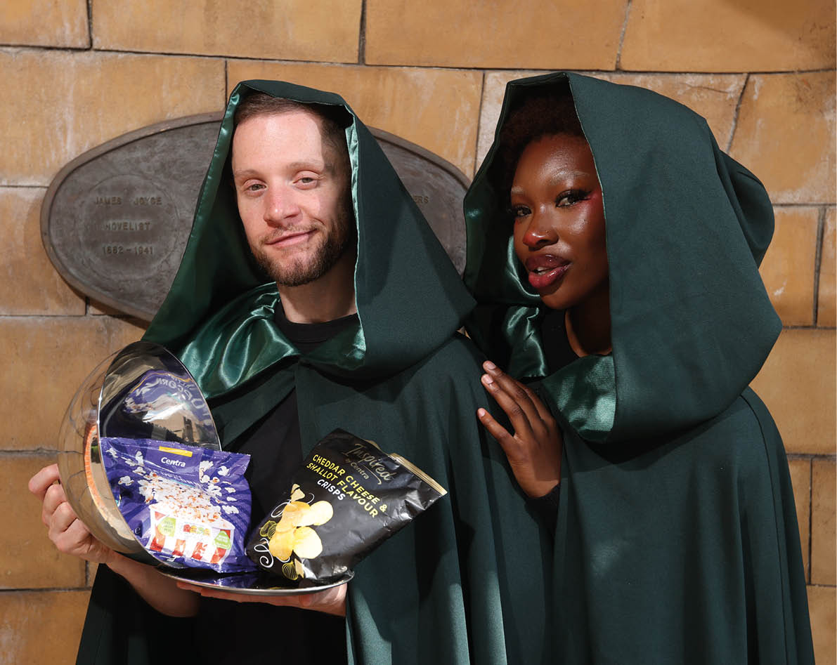 Two people wearing dark green cloaks with hoods pose together while holding a tray with various packaged snacks in front of a stone wall. Description generated by AI