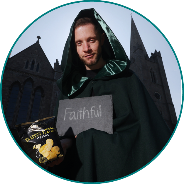 A person wearing a dark green hooded cloak stands outdoors near a stone building holding a sign that reads “Faithful." Description generated by AI