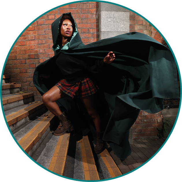 A person wearing a dark green flowing cloak with a hood and a red plaid skirt is striking a dynamic pose against a brick and stone wall background. Description generated by AI