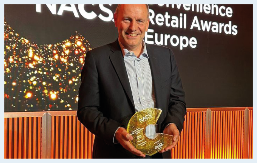 A person in a suit is smiling and holding a golden award shaped like the letter C at a retail awards event with a lit backdrop. Description generated by AI