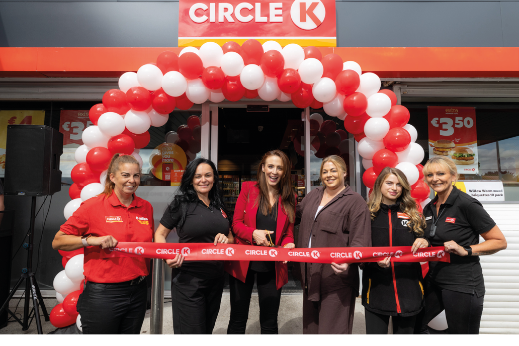 Six adults stand smiling in front of a Circle K store under a red and white balloon arch while holding a red ribbon for a ribbon-cutting ceremony. Description generated by AI