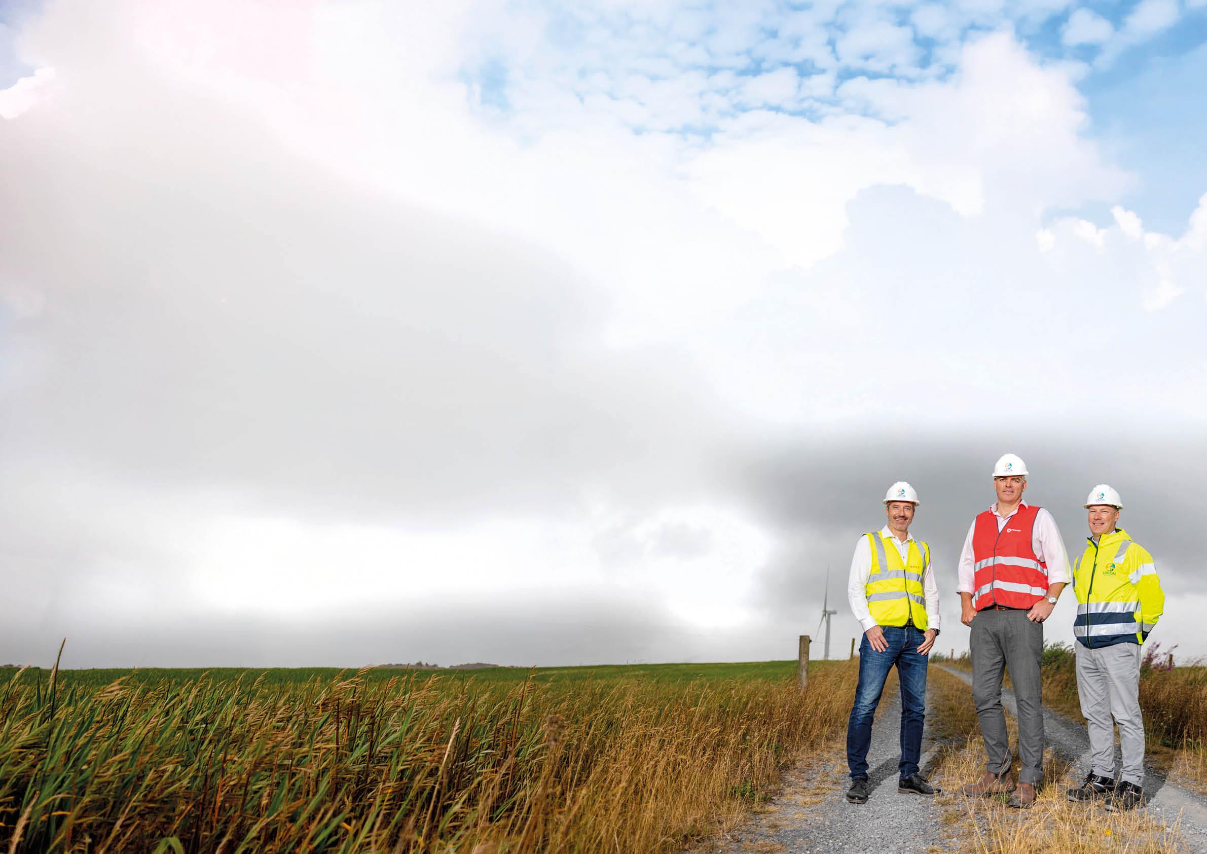 Three adults wearing safety helmets and high-visibility vests stand on a gravel path next to tall grass under a cloudy sky. Description generated by AI