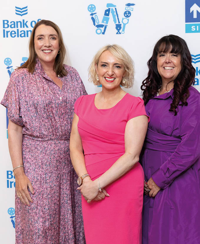 Three adults dressed in colorful dresses stand smiling in front of a backdrop with logos including Bank of Ireland. Description generated by AI