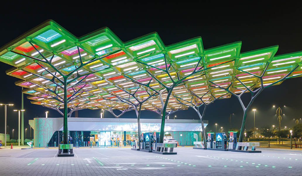 A brightly lit modern gas station at night features a colorful, geometric canopy with green, red, and white illuminated panels above the fuel pumps and a small building in the background. Description generated by AI