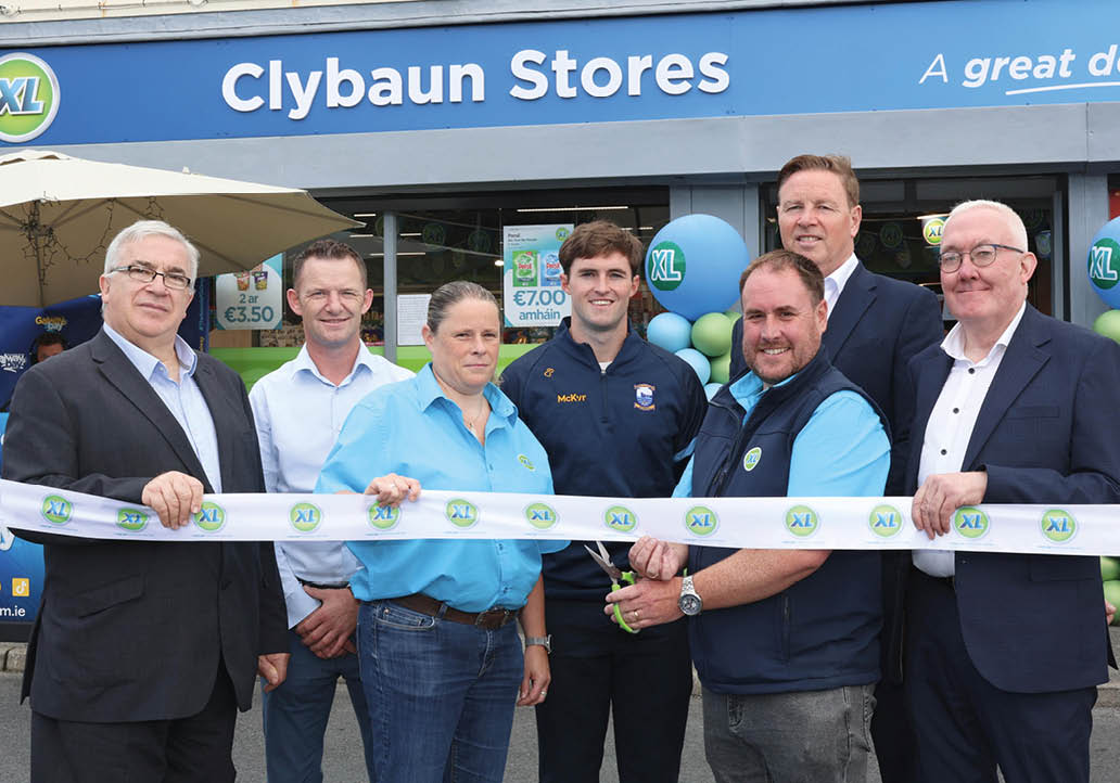 Seven adults stand in front of a store named Clybaun Stores holding a ribbon for a ribbon-cutting event with balloons and promotional signs visible in the background. Description generated by AI