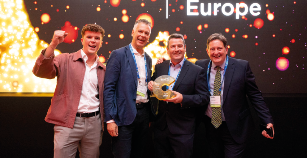 Four adults dressed in business and casual attire celebrate on stage with one holding a trophy and a backdrop displaying the word Europe. Description generated by AI