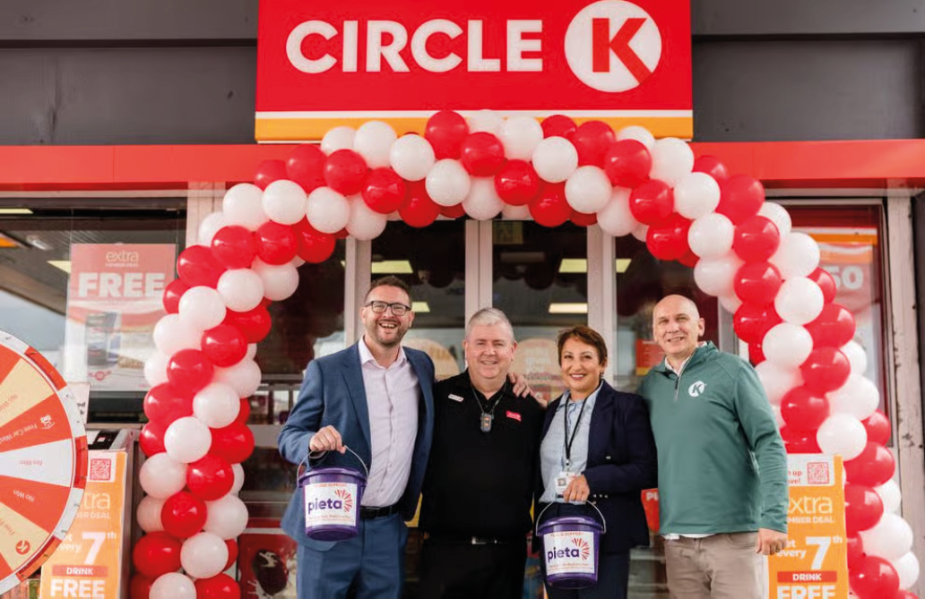 Four adults smiling and holding buckets stand under a red and white balloon arch in front of a Circle K store. Description generated by AI