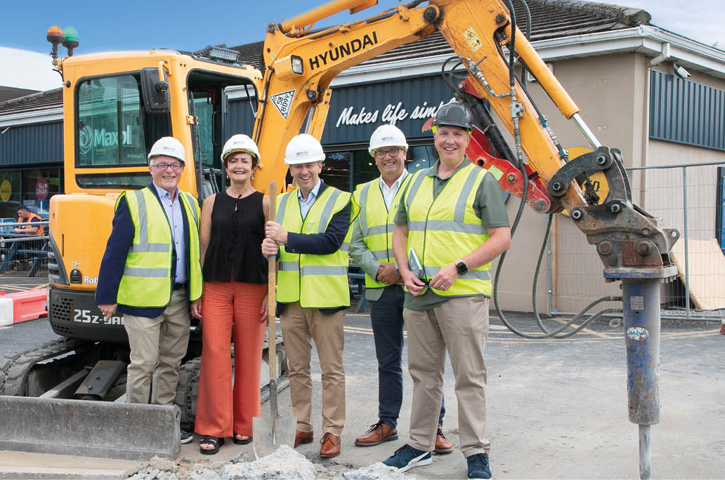 Five adults wearing safety helmets and high-visibility vests stand smiling in front of construction machinery at a worksite, with one person holding a shovel. Description generated by AI
