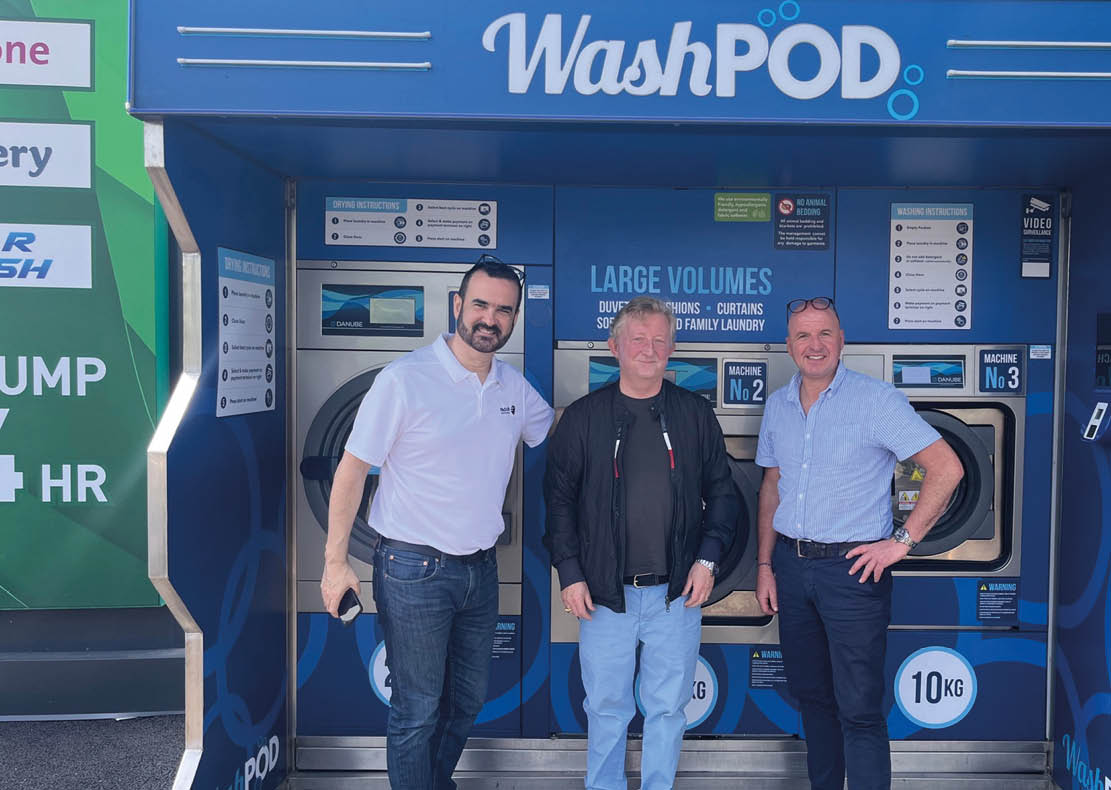 Three adults stand smiling in front of a self-service laundry machine area labeled WashPOD, with two of them wearing casual shirts and jeans and one wearing a black jacket and light blue pants. Description generated by AI