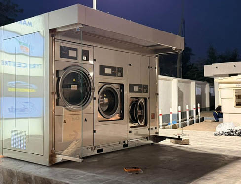 A modern outdoor laundry station with three large stainless steel washing machines is set on a tiled platform at dusk with a person sitting nearby. Description generated by AI