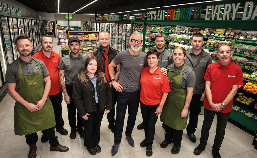 A group of adults wearing store uniforms and aprons stand together smiling inside a grocery store aisle filled with fresh produce and packaged goods. Description generated by AI