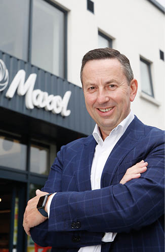 A person wearing a blue suit jacket and white shirt is standing with arms crossed and smiling in front of a building with a sign that reads “Maxol." Description generated by AI