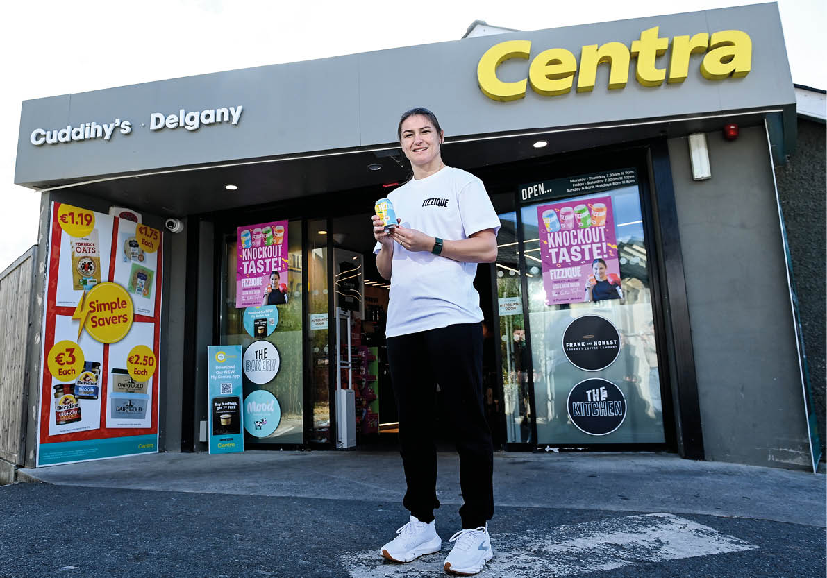 A person wearing a white t-shirt and black pants is standing outside a Centra convenience store holding a small item and smiling. Description generated by AI