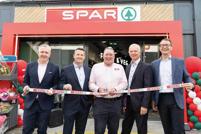 Five adults in business attire stand smiling in front of a SPAR store holding a branded ribbon for a ribbon-cutting ceremony. Description generated by AI