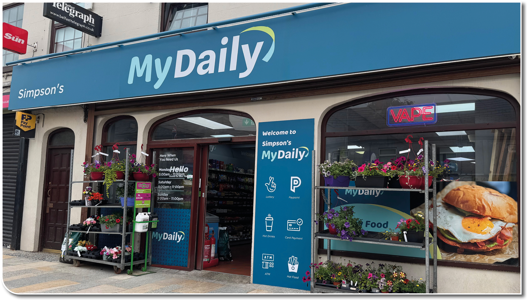 A storefront with a blue sign reading “MyDaily" and flower racks outside displays a neon "OPEN" sign and a large image of a breakfast sandwich in the window. Description generated by AI