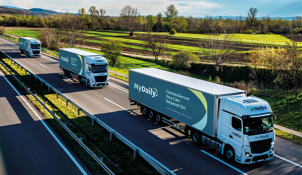 Three white trucks with trailers are driving in a line on a divided highway surrounded by green fields under a partly cloudy sky. Description generated by AI