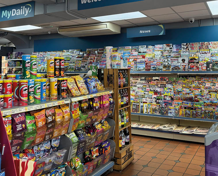 A brightly lit newsagent store interior with shelves stocked with snacks, magazines, and newspapers under signs reading “Welcome to Simpson's Newsagent" and "Mydaily." Description generated by AI