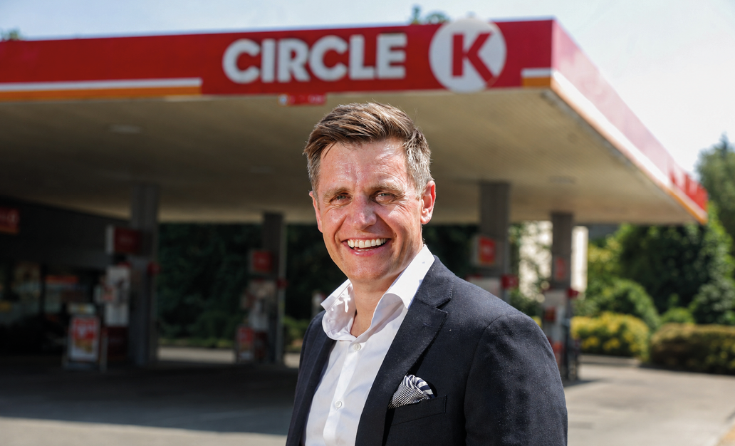 A person wearing a dark blazer and white shirt is smiling and standing in front of a Circle K gas station. Description generated by AI