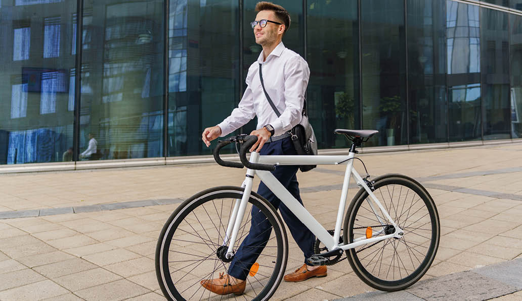 A professional man walks his bicycle beside a modern glass office building, exuding confidence and style.